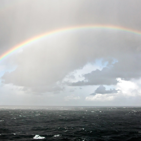 Black and white ocean and cloudy sky above it. A rainbow stretches from the left side across to right side of photo.