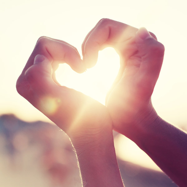 Two hands coming together to form a heart, with sunlight beaming through the heart from the background.
