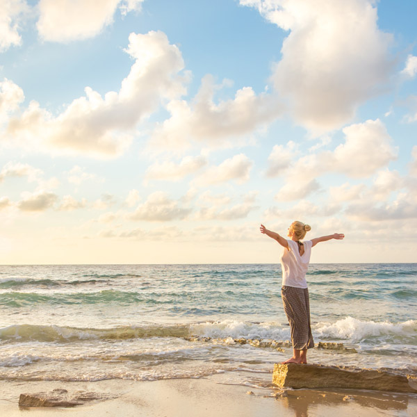 A woman standing on the shore in front of the ocean. The upper portion of the sky is filled with blue sky and clouds.