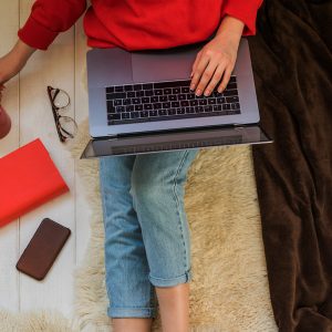 A woman's body in a red sweater laying on a bench with her laptop, phone, a red book, and glasses.