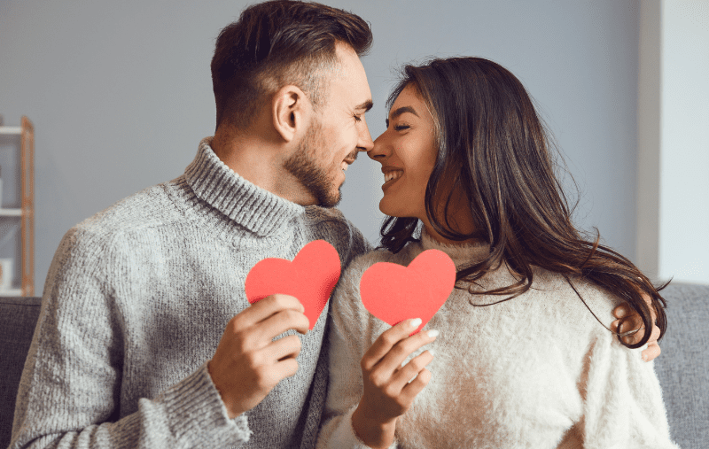A happy couple facing each other, smiling, with their noses touching while each are holding a red heart
