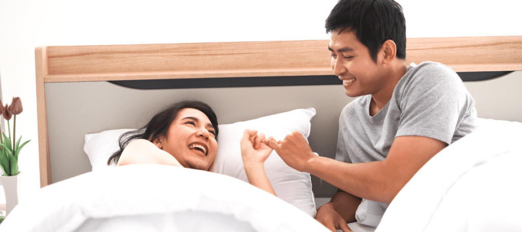 A man and a woman laying in bed together, laughing, with their pinky fingers intertwined.